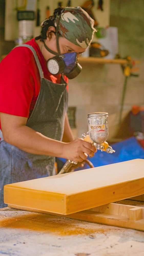 Aplicación de acabado con pistola sobre una pieza de madera, con mascarilla de protección